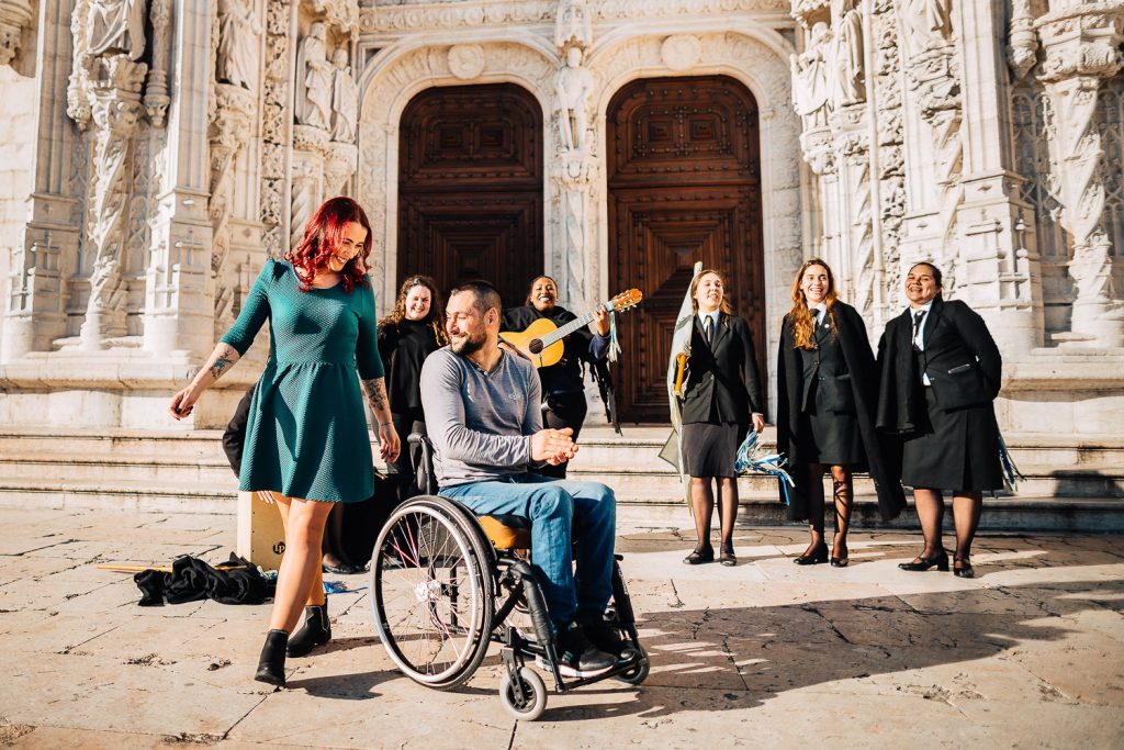 noiva dançando e noivo na cadeira de rodas batendo palmas com algumas estudantes cantando e tocando em frente ao Mosteiro dos Jeronimos