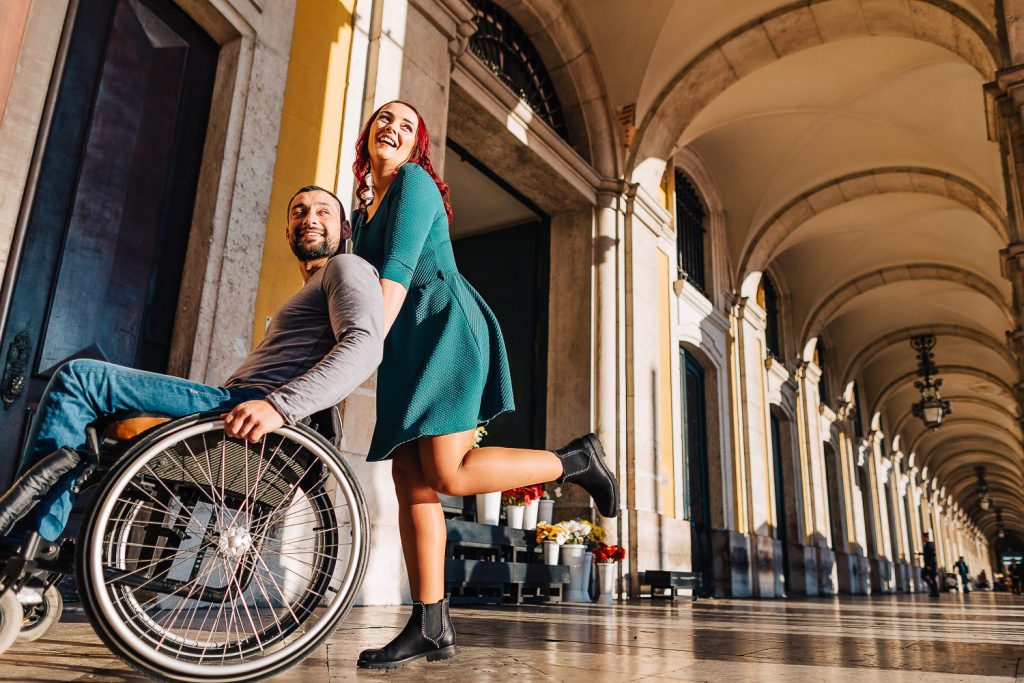 Noiva empurrando a cadeira de rodas e os dois sorrindo no arco da rua augusta em Lisboa