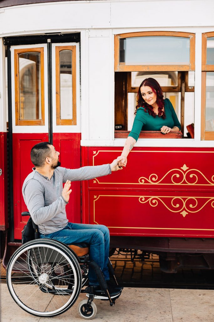 Casal no bondinho vermelho em Lisboa, com a noiva na janela dentro do bondinho e o noivo segurando as mão do lado de fora na cadeira de rodas