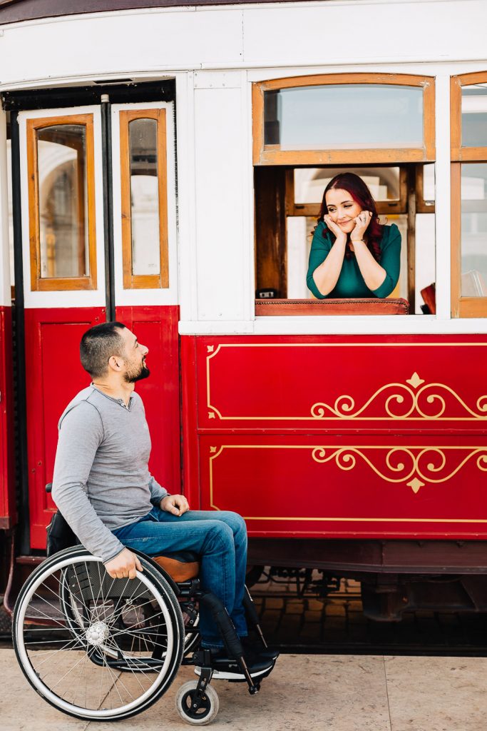 Casal no bondinho vermelho em Lisboa, com a noiva na janela dentro do bondinho e o noivo admirando do lado de fora na cadeira de rodas